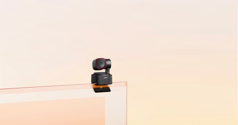 A small black camera with a red trim sits atop a thin computer monitor against a gradient beige background. The camera has a circular lens and a square-shaped body, suggesting it's a webcam designed for video conferencing or streaming.