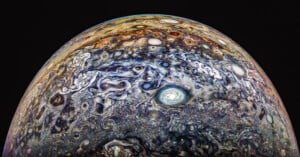 Colorful and swirling cloud patterns on Jupiter's atmosphere are shown, featuring a prominent white circular storm in the center, with various shades of blue, brown, and orange throughout the gaseous landscape under a dark sky.