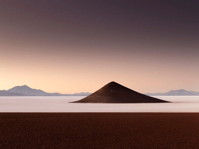 A lone conical hill rises from a flat, expansive salt plain, surrounded by a vast, arid landscape. Distant mountains are faintly visible under a gradient sky transitioning from warm tones to a dusky shade.