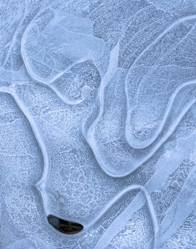 Abstract close-up of icy formations with smooth, curving lines and a frosty texture. A dark, leaf-like shape is embedded in the bottom section. The overall color palette is a mix of light blues and whites, creating an ethereal, wintry feel.