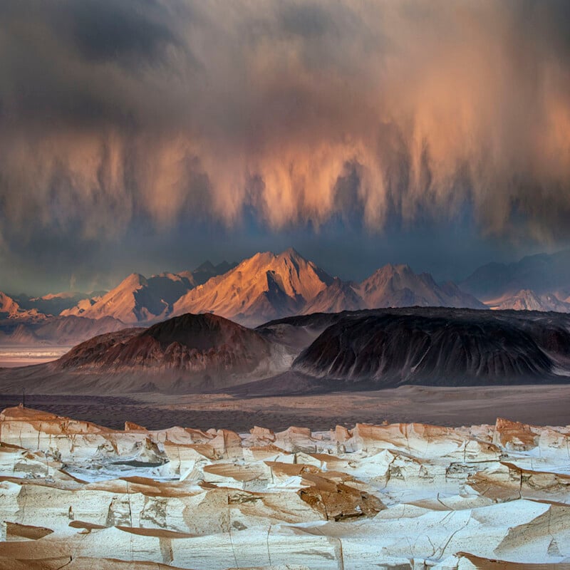 Breathtaking scene of a rugged desert landscape under a dramatic sky. Dark clouds hang over distant mountains, illuminated by soft sunlight. The foreground features cracked earth and rock formations, creating an intricate natural pattern.