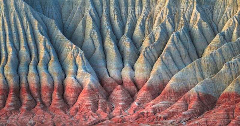 Eroded, multicolored geological formations create striking natural patterns on a rugged cliff face. Vertical striations in shades of gray, tan, and reddish-brown dominate the scene, resembling draped fabric across the landscape.