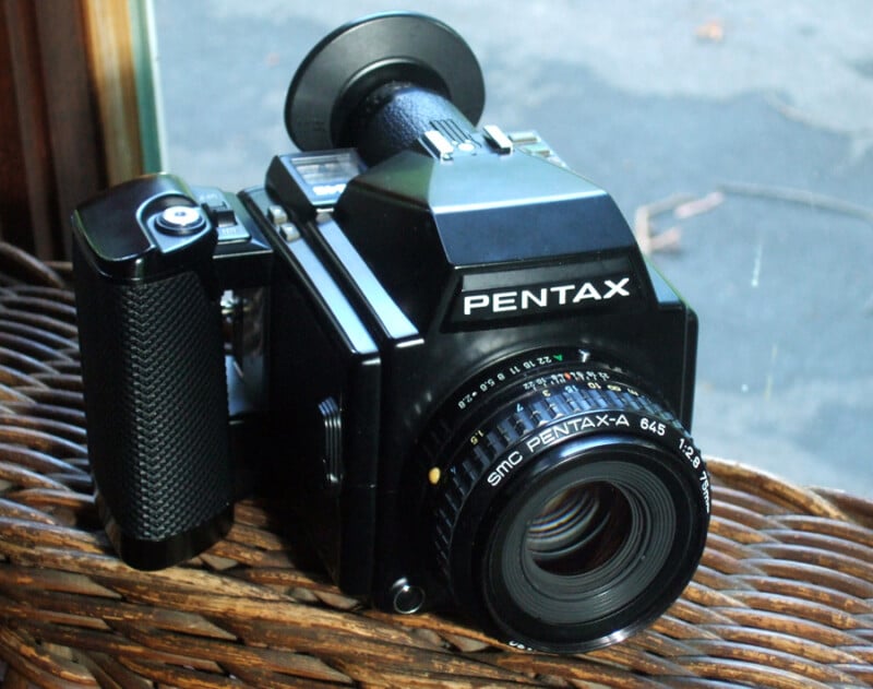 A vintage black Pentax medium format camera with a grip and lens, resting on a woven surface by a window, capturing natural light.