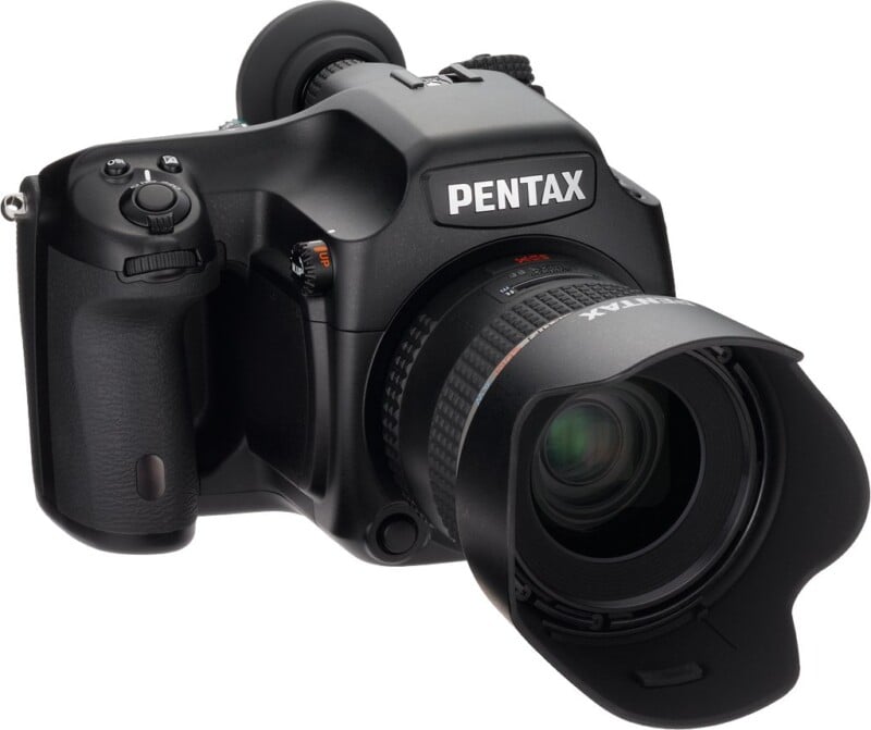 A black Pentax digital camera with a large lens and lens hood, displayed at an angle. The camera has a textured grip and various buttons for controls. The Pentax logo is visible on the body of the camera.