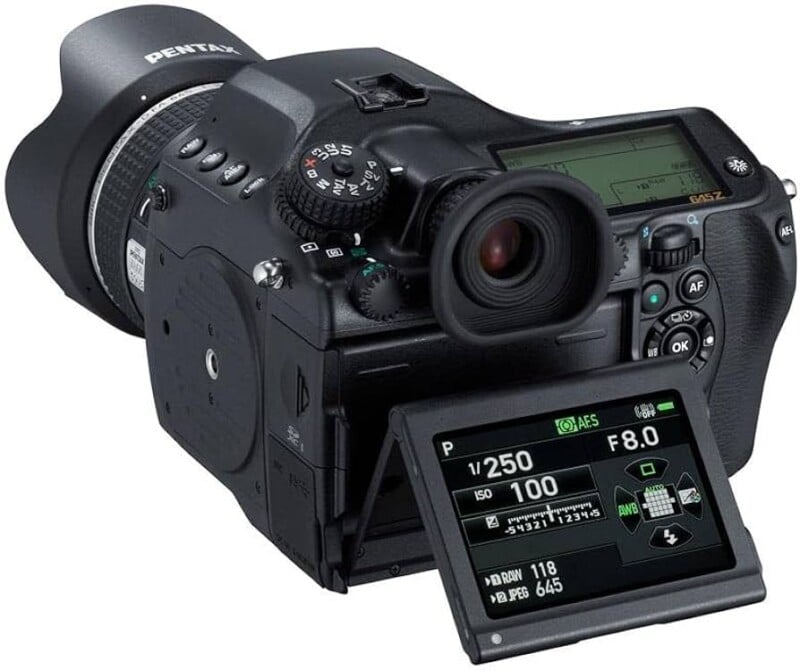 A black Pentax digital camera with a large lens and a tilting LCD screen displaying camera settings, including shutter speed, aperture, and ISO. The camera has multiple buttons and dials for manual adjustments.