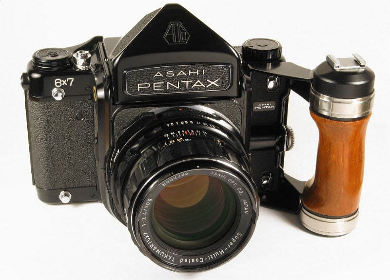 A black Asahi Pentax 6x7 film camera with a wooden hand grip is facing forward. The lens displays details like aperture and focal length markings. The camera has a robust, vintage appearance.