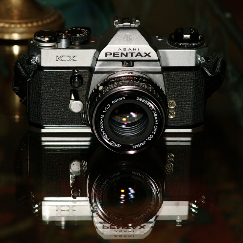 A vintage Asahi Pentax KX film camera is placed on a reflective surface, highlighting its well-crafted design. The camera's various controls and branding are visible, showcasing its classic aesthetic.