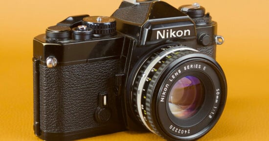 A vintage Nikon film camera with a 50mm f/1.8 lens is displayed against an orange background. The camera features a black body with manual controls and a classic design.