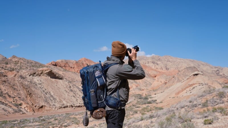 A person wearing a beanie and a backpack stands in a desert landscape, taking photos with a camera. Rugged, multicolored hills and a clear blue sky are in the background.