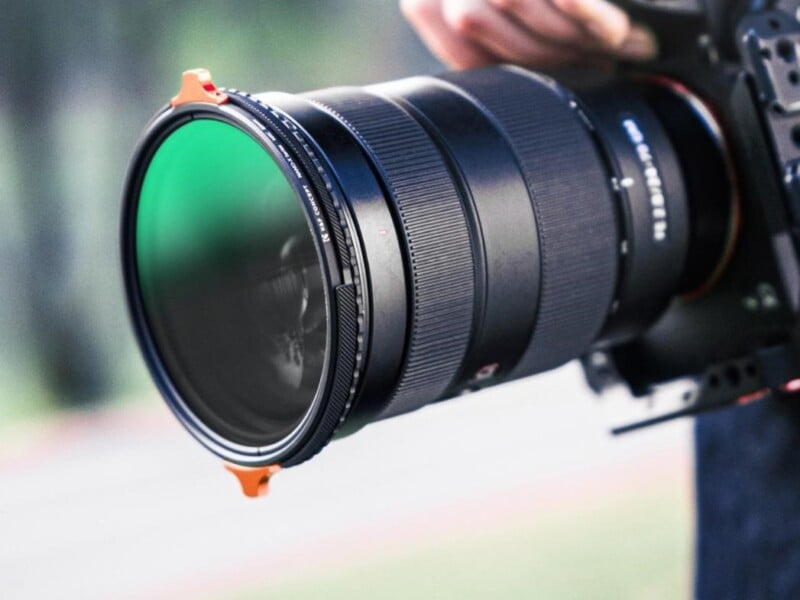 Close-up of a large camera lens with a green-tinted filter attached. The lens is held by a person, partially visible, against a blurred outdoor background, suggesting a photography session.