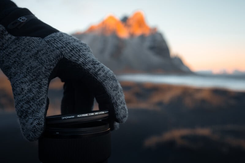 A gloved hand holds a camera lens in the foreground. In the background, a mountainous landscape is softly illuminated by the warm light of the setting sun, creating a serene and picturesque scene.