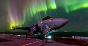 A fighter jet is parked on an aircraft carrier deck under a vivid display of the northern lights. The night sky is filled with colorful waves of green and yellow auroras, enhancing the dramatic scene.