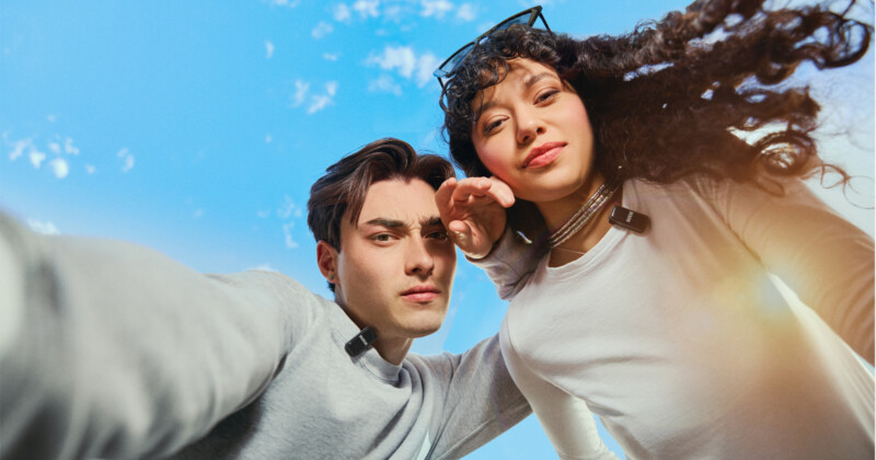 Two people in casual clothing looking down at the camera against a bright blue sky. The person on the left has short brown hair, and the person on the right has long curly hair and sunglasses on their head. Both appear to be taking a selfie.