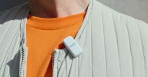 A person wearing a light gray jacket and an orange shirt has a small white RODE microphone clipped to their collar. The image focuses on the microphone and part of the upper torso.