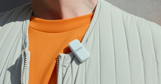 A person wearing a light gray jacket and an orange shirt has a small white RODE microphone clipped to their collar. The image focuses on the microphone and part of the upper torso.