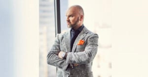 A man with a beard stands with his arms crossed, gazing out of a window. He wears a grey tweed jacket, a dark shirt, and an orange pocket square, with a watch on his wrist. The background shows blurred city buildings.