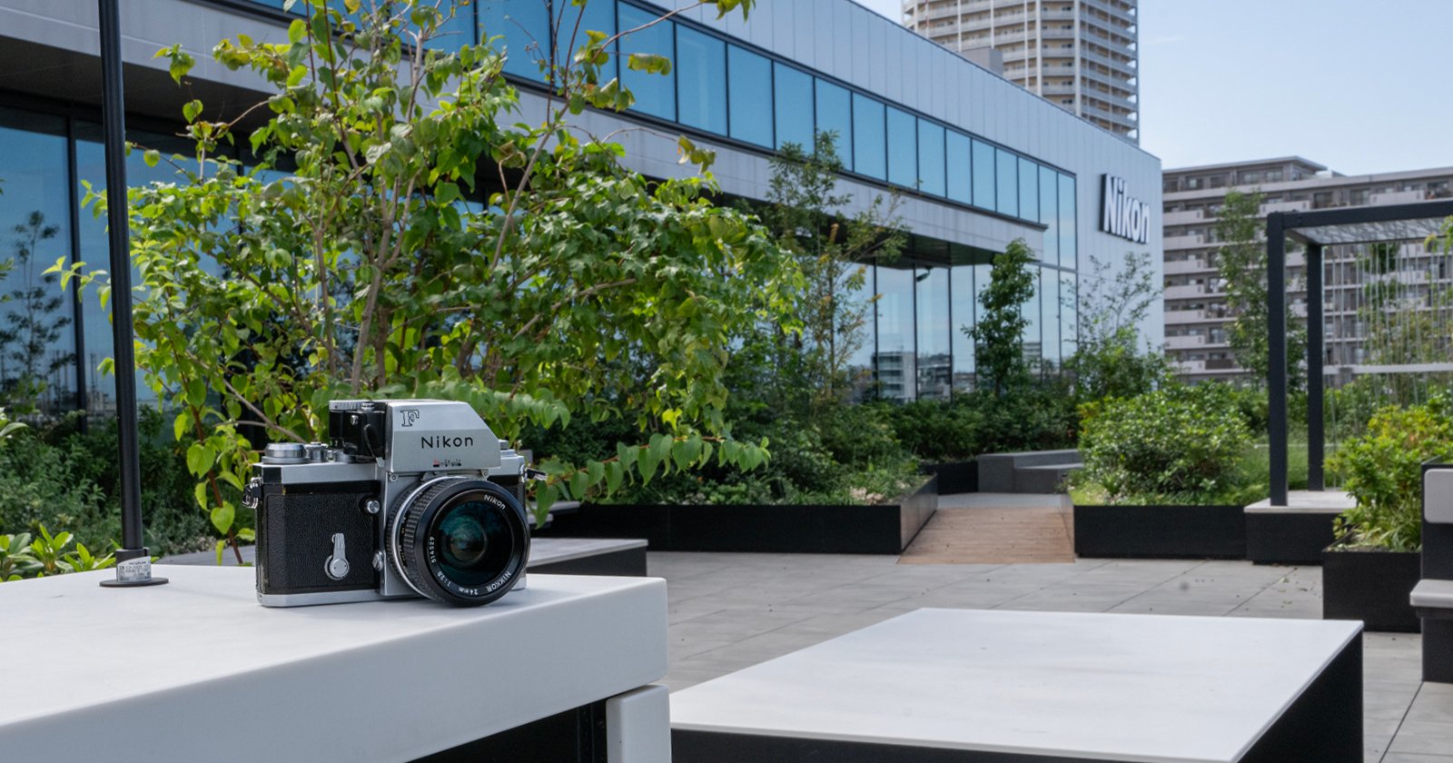 A Nikon camera sits on a white table outdoors with a modern building and greenery in the background. The building has large glass windows and a visible Nikon logo.