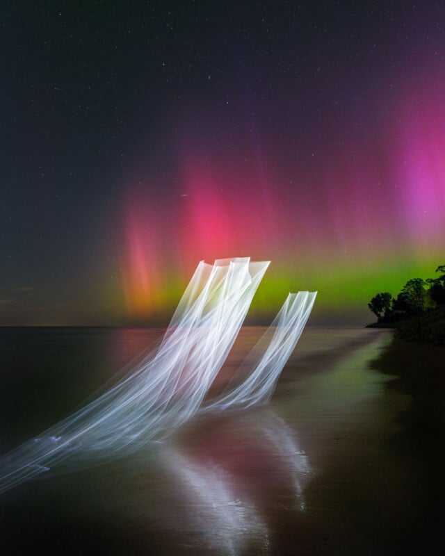 A vibrant Northern Lights display with ribbons of green and pink hues illuminates the night sky. Wispy white patterns resemble waves on a calm shoreline, blending into the reflections on the water's surface. Trees silhouette on the right horizon.