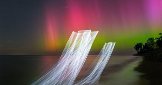 Colorful auroras in the night sky with pink, red, and green lights swirling above a dark coastline. A ghostly, elongated light effect stretches from the water towards the sky, creating an ethereal scene.