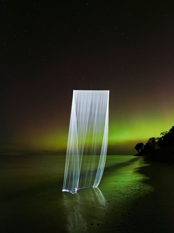 A long exposure photograph of thin, white light streams forming a rectangular shape over a dark beach. The Aurora Borealis glows green in the background sky, reflecting on the water. Silhouetted trees line the shoreline.