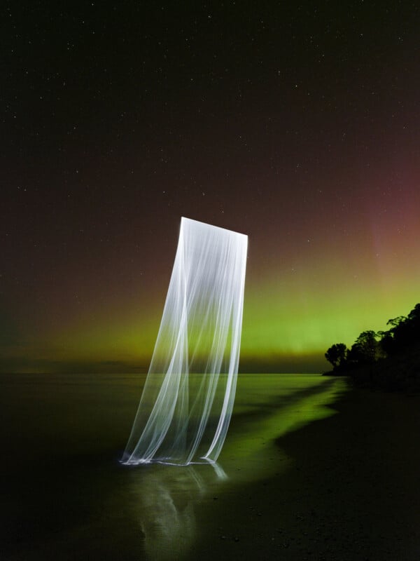 A long-exposure photograph shows white light trails forming a vertical sheet against a backdrop of the aurora borealis and a starry night sky, with silhouettes of trees along the shoreline.