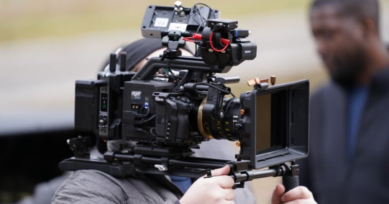 A person holds a large professional video camera equipped with various attachments and cables. The equipment is detailed and prominent, with a blurred background and another individual slightly visible out of focus.