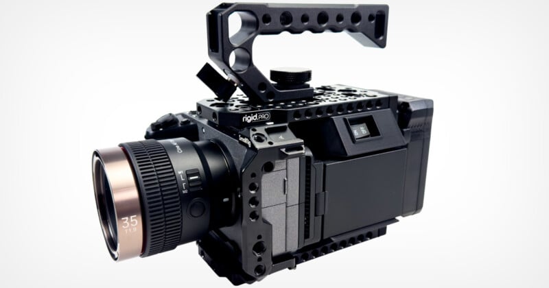 A professional cinema camera with a detachable handle, matte black finish, and a 35mm T1.9 lens is shown. The camera is encased in a sturdy, perforated metal frame for added protection and mounting versatility.