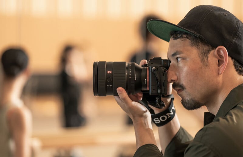 A man wearing a cap holds a professional camera close to his face, focusing intently. The background is softly blurred, with indistinct figures and a neutral indoor setting.