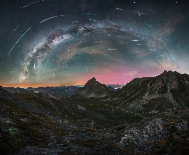 A stunning night sky over a mountainous landscape, featuring a majestic arc of the Milky Way and vibrant meteor streaks. The foreground showcases rugged terrain and a winding path through green hills under the starry sky.