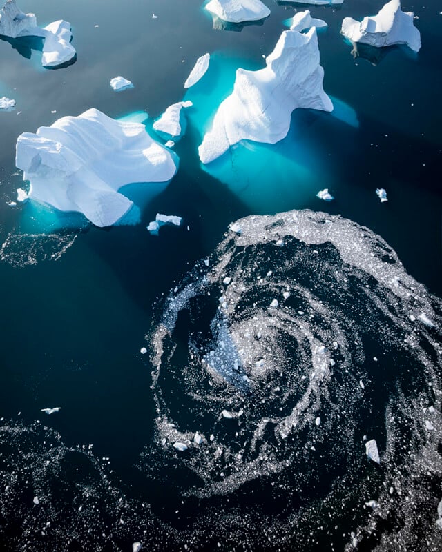 Aerial view of icebergs floating in dark blue water. White ice contrasts with the deep turquoise of the submerged ice. A swirling pattern of ice crystals creates a spiral effect on the water's surface near the icebergs.