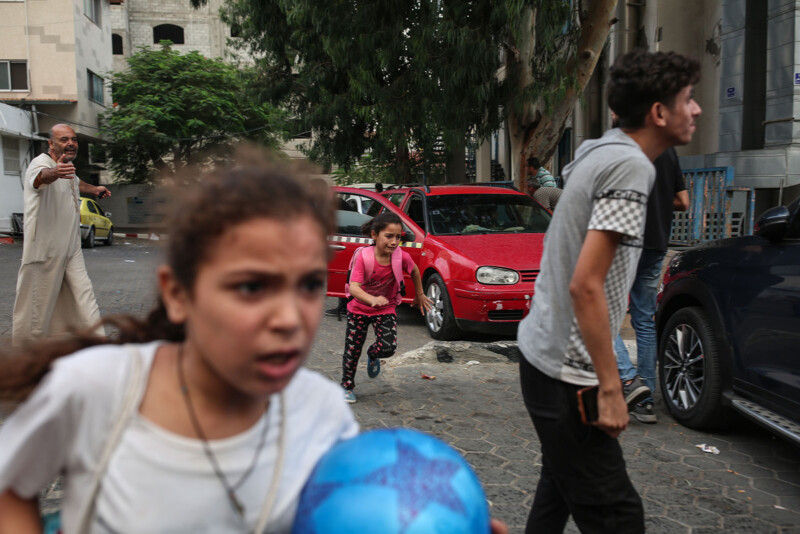 Children running on a street with concerned expressions, one holding a blue ball. A man in the background gestures, while parked cars and trees are visible. The scene suggests urgency or alarm.