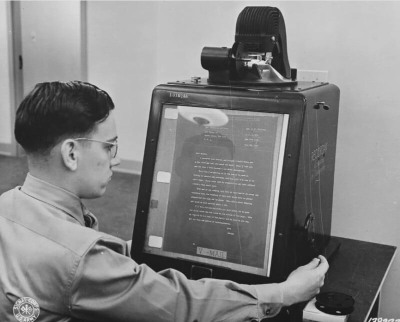 A man operates a vintage reading machine for the visually impaired. The device displays magnified text on a screen. He adjusts knobs on the side while seated in a room, wearing glasses and a uniform shirt.