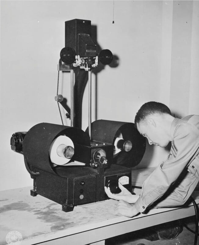 A man in a lab coat operates a vintage machine with two large rotating drums and various levers on a table. The setting appears to be a laboratory or industrial environment.