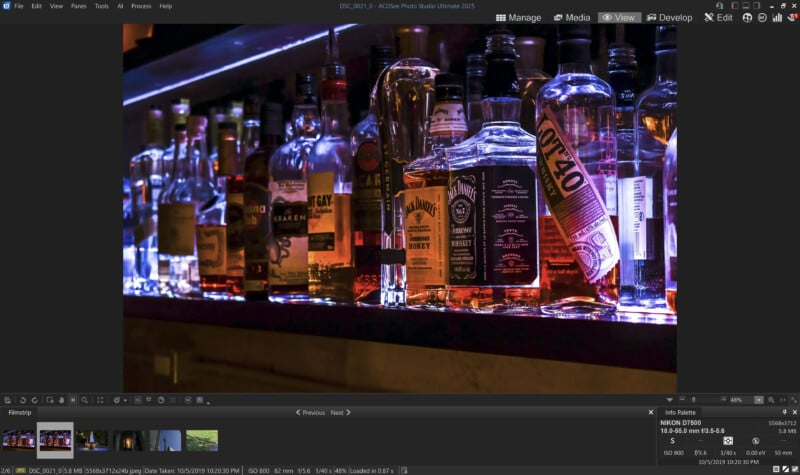 A dimly lit bar shelf displays various liquor bottles, including whiskey and rum, set against a backdrop of blue and purple neon lights. The reflective bar surface adds a sleek, modern touch to the scene.