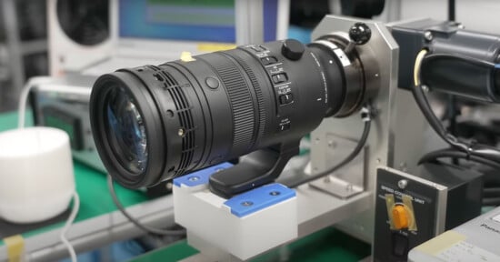 Close-up of a large camera lens mounted on a testing rig in a laboratory setting. The lens has various control switches and is positioned on a green surface with electronic equipment visible in the background.