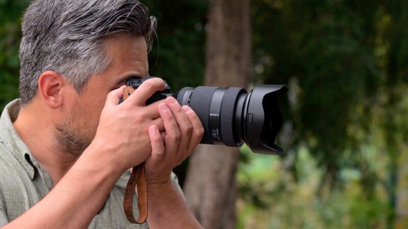 A person with gray hair holds a professional camera up to their eye, focusing on capturing an image. The background is blurred greenery, suggesting an outdoor location. The individual wears a casual, light-colored shirt.