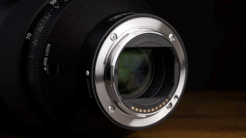 Close-up of a camera lens mount with a metallic ring and electronic contacts. The lens is partially shadowed, and the focus is on the intricate details of the mount.