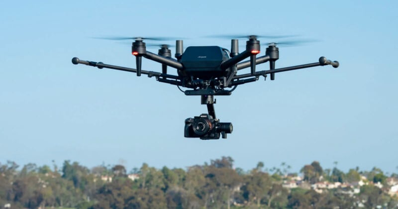 A quadcopter drone equipped with a camera hovers in the sky. The background shows a blurred landscape of trees and buildings under a clear blue sky.