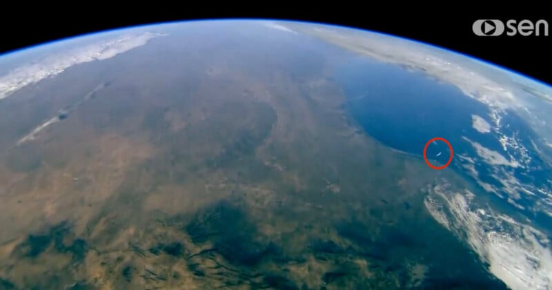 View of Earth from space showing continents and ocean. A red circle highlights a small object near the coast. The horizon curves with the planet's surface, and the Sen logo is visible in the corner.