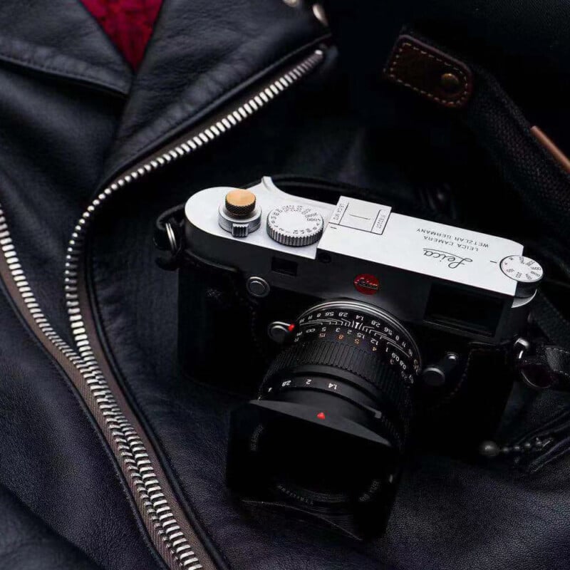 A Leica camera resting on a partially unzipped black leather jacket. The camera is positioned with its top view visible, showcasing its dials and lens. The background includes hints of a dark bag and red lining.