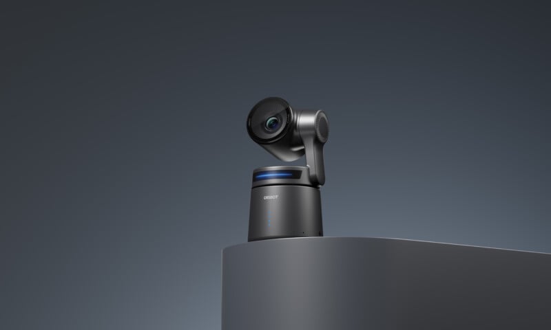 A sleek, modern camera with a rotating head and blue accent, placed on a curved, gray surface against a dark background.