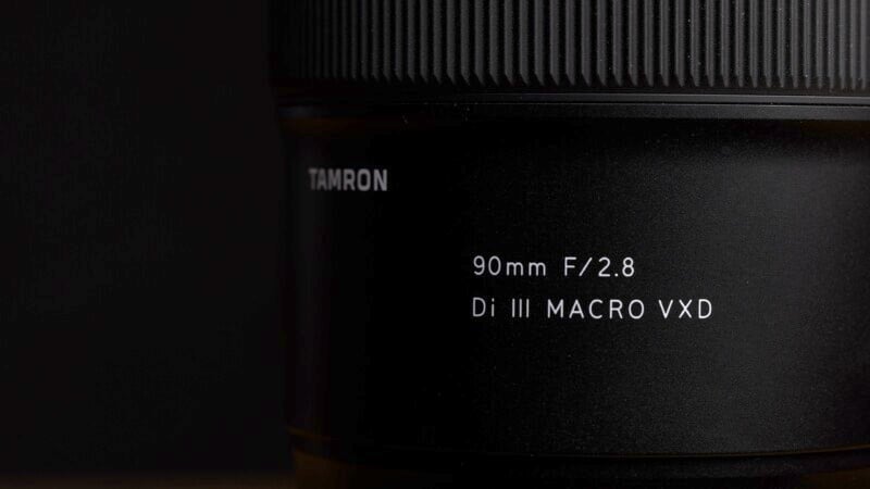 Close-up of a black camera lens with engraved text showing "Tamron 90mm F/2.8 Di III MACRO VXD" on a dark background. The focus is on the text, partially illuminated by light.