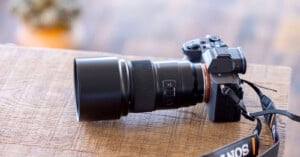 A black camera with a large telephoto lens rests on a wooden table. The camera strap, partially visible, is branded with "SONY". The background is softly blurred, highlighting the camera as the focal point.