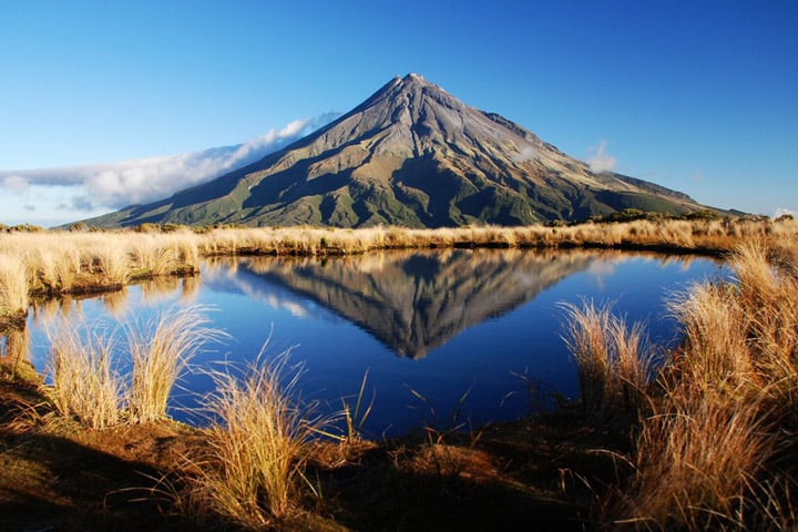 A serene landscape of a volcano with a conical peak against a clear blue sky. The volcano is reflected in a still pond surrounded by dry golden grass, creating a symmetrical, picturesque scene.