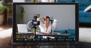 A woman sits at a table facing a camera and a smartphone on a tripod. She is looking at her phone screen, adjusting her hair. The table holds a ring light, laptop, makeup, and other filming equipment. The setting looks like a home studio.