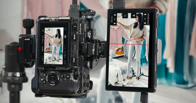 A camera and a smartphone on stands are capturing a woman in a cozy room. She is standing with her arm extended, wearing a beige sweater and jeans. Behind her, there are hanging clothes and a soft, ambient setup.