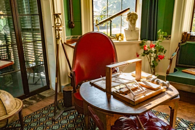 A vintage study room with a red chair and wooden desk. On the desk is a drafting tool. A telescope and bust decorate the room, with green walls and a large window letting in natural light. Red flowers add a pop of color.