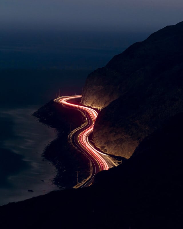 La nuit, une route côtière sinueuse montre des traînées de lumière rouge et blanche provenant des véhicules. La route est bordée d’un côté par des falaises sombres et de l’autre par l’océan, créant une vue spectaculaire et panoramique.