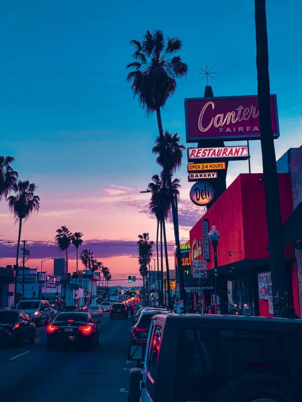 Une scène de rue animée au coucher du soleil avec des palmiers bordant la route. Les voitures sont visibles au premier plan. Les enseignes au néon d'un restaurant, d'une épicerie fine et d'une boulangerie brillent sur le ciel rose et violet, ajoutant une atmosphère vibrante.