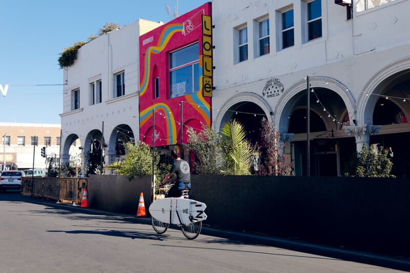 Une personne faisant du vélo avec une planche de surf attachée passe devant des bâtiments colorés dans une rue ensoleillée.